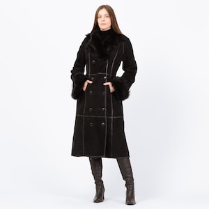 May include: A long, black coat with a double-breasted design. The coat features a faux fur collar and cuffs, with dark brown stitching and buttons. The coat is knee-length and worn with dark brown boots.
