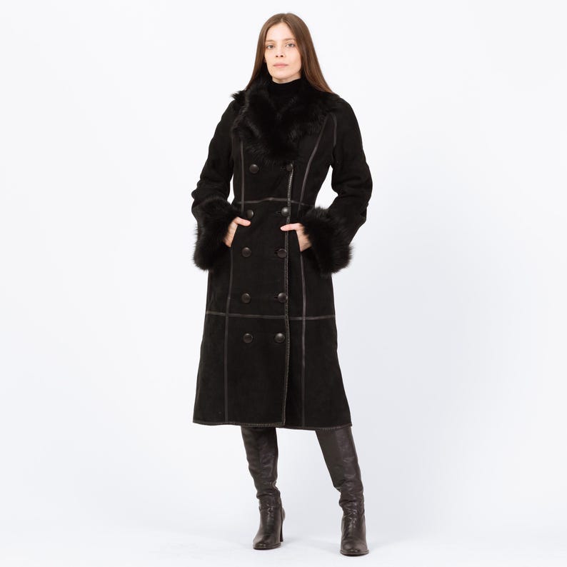 May include: A long, black coat with a double-breasted design. The coat features a faux fur collar and cuffs, with dark brown stitching and buttons. The coat is knee-length and worn with dark brown boots.