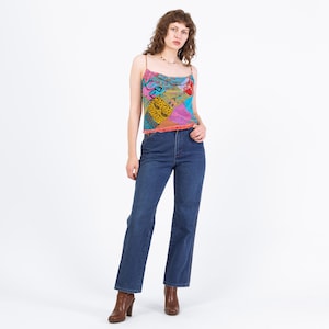 May include: A woman wearing a colorful patchwork print camisole top with spaghetti straps and dark blue jeans. The top features a variety of patterns and colors, including pink, yellow, and blue. She is also wearing brown heeled boots.