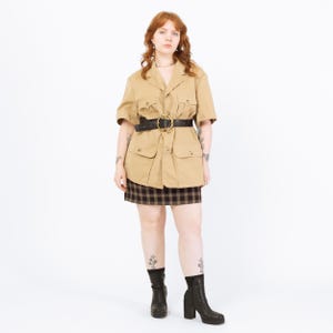 Puede incluir: Chaqueta de estilo safari color beige con mangas cortas, cuatro bolsillos delanteros y cinturón negro con hebilla dorada. La chaqueta se combina con una minifalda a cuadros y botines negros con tacón. El conjunto es una mezcla de vintage y moderno.