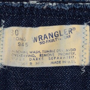 K&ouml;nnte beinhalten: Nahaufnahme eines Wrangler Jeans-Etiketts. Das cremefarbene Etikett tr&auml;gt den blauen Schriftzug "WRANGLER" und "NO-FAULT JEAN". Das Etikett enth&auml;lt auch Waschanweisungen und "MADE IN U.S.A."