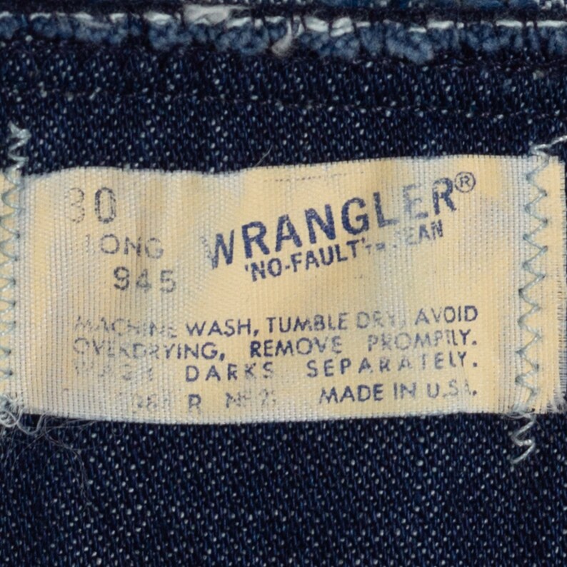 K&ouml;nnte beinhalten: Nahaufnahme eines Wrangler Jeans-Etiketts. Das cremefarbene Etikett tr&auml;gt den blauen Schriftzug "WRANGLER" und "NO-FAULT JEAN". Das Etikett enth&auml;lt auch Waschanweisungen und "MADE IN U.S.A."