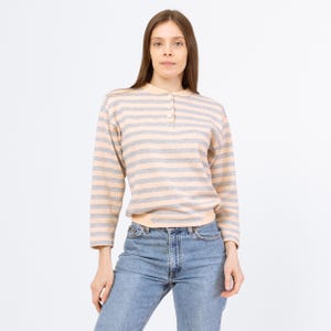 May include: A long-sleeved striped shirt with a button-up collar. The shirt is a light pink and gray color. The model is wearing blue jeans.