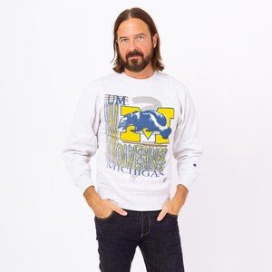 Könnte beinhalten: Ein hellgraues Sweatshirt mit einer großen Grafik mit einem blau-weißen Bären, dem Wort "Wolverines" in Gelb und dem Text "Michigan". Das Sweatshirt hat lange Ärmel und einen Rundhalsausschnitt.