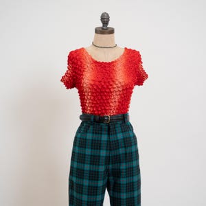 May include: A short-sleeved top with a textured, crinkled surface that transitions from a bright red at the sleeves to a lighter orange-red in the center. It is paired with high-waisted, dark green and black plaid trousers and a black belt.