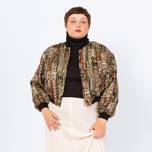 May include: A patterned bomber jacket in shades of green, brown, and red. The jacket has a black collar and cuffs. The model is wearing a black turtleneck and a cream-colored skirt. The jacket's design features geometric and abstract shapes.
