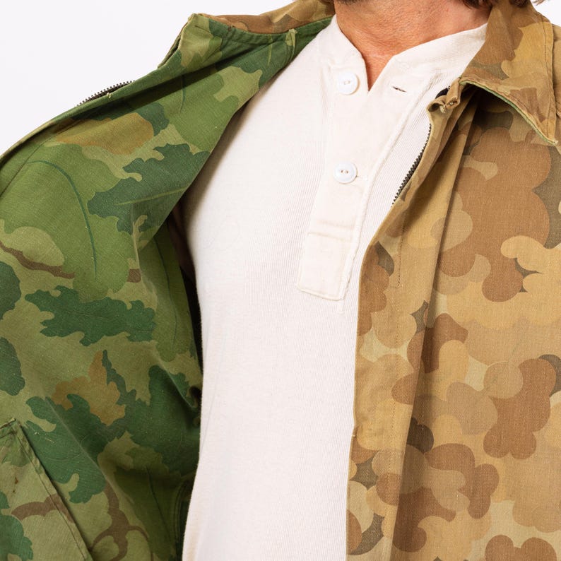May include: A reversible camouflage jacket with a green and brown pattern on one side and a tan and brown pattern on the other. The jacket is open, revealing a white Henley shirt underneath.
