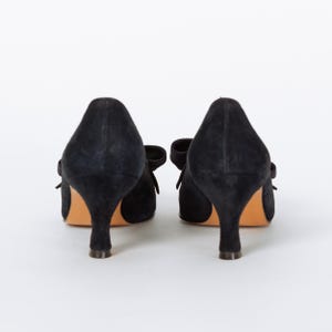 Puede incluir: Un par de tacones negros de ante con peque&ntilde;os lazos y un tac&oacute;n bajo. Los zapatos est&aacute;n sobre un fondo blanco, mostrando su elegante dise&ntilde;o. Los tacones tienen un estilo cl&aacute;sico y atemporal.