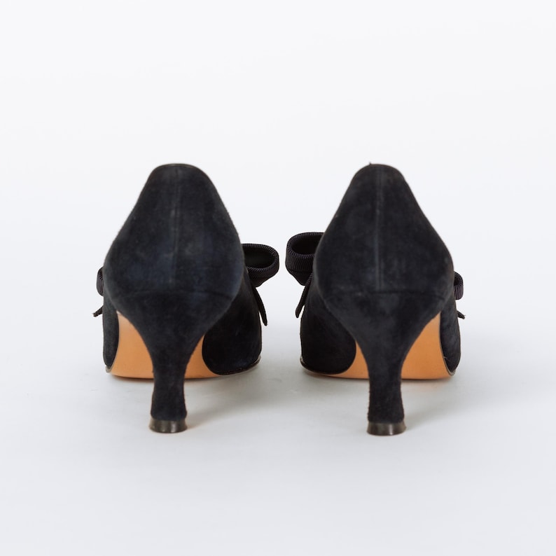 Puede incluir: Un par de tacones negros de ante con peque&ntilde;os lazos y un tac&oacute;n bajo. Los zapatos est&aacute;n sobre un fondo blanco, mostrando su elegante dise&ntilde;o. Los tacones tienen un estilo cl&aacute;sico y atemporal.