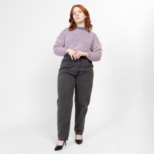 Peut inclure: Une femme portant un pull violet clair et un jean gris foncé. Elle est debout, les mains jointes devant elle.