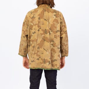 May include: A tan and brown camouflage pattern jacket with long sleeves. The jacket has a green trim along the bottom and sleeve cuffs. The person is wearing dark blue jeans.