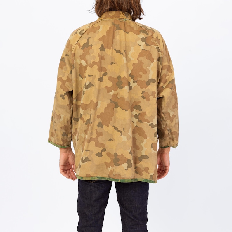 May include: A tan and brown camouflage pattern jacket with long sleeves. The jacket has a green trim along the bottom and sleeve cuffs. The person is wearing dark blue jeans.
