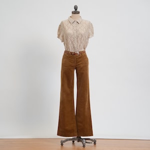 May include: A beige short-sleeved lace blouse with a collar and front button closure paired with brown corduroy wide-leg trousers featuring a brown leather belt with a silver buckle, displayed on a mannequin against a plain background.