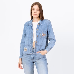 May include: Light blue denim jacket with long sleeves, a collared neckline, and wooden buttons. The jacket features embroidered patches on the pocket and lower left side. The model is wearing a white top and light blue jeans.