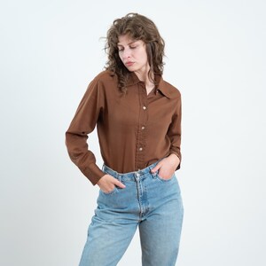 May include: A long-sleeved brown button-down shirt with a pointed collar and a western-style yoke detail across the chest, paired with light blue denim jeans. The shirt features a front button placket and long sleeves with buttoned cuffs.
