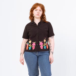 Może przedstawiać: Ciemnobrązowa koszula zapinana na guziki z krótkim rękawem z haftowanymi kwiatami w donicach wzdłuż dolnego obszycia. Kwiaty są różowe, żółte i białe, z zielonymi liśćmi i kolorowymi donicami. Koszula jest połączona z niebieskimi dżinsami.
