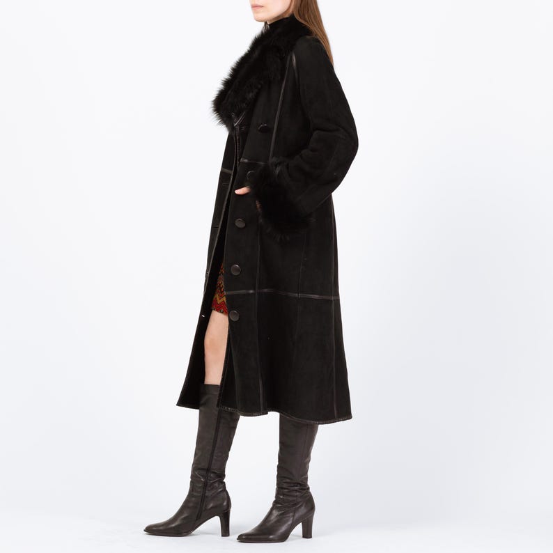 May include: A long, black coat with a fur collar and cuffs. The coat has a button-down front and is worn with black knee-high boots. The coat appears to be made of suede or a similar material. The overall style is classic and elegant.