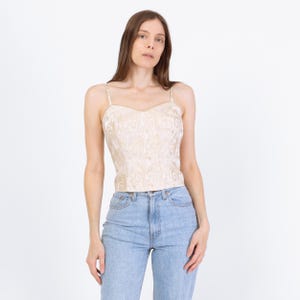 May include: A cream-colored bustier top with a subtle, swirling pattern. The top features thin spaghetti straps and a sweetheart neckline. The garment is paired with light blue denim jeans. The overall style is vintage-inspired.