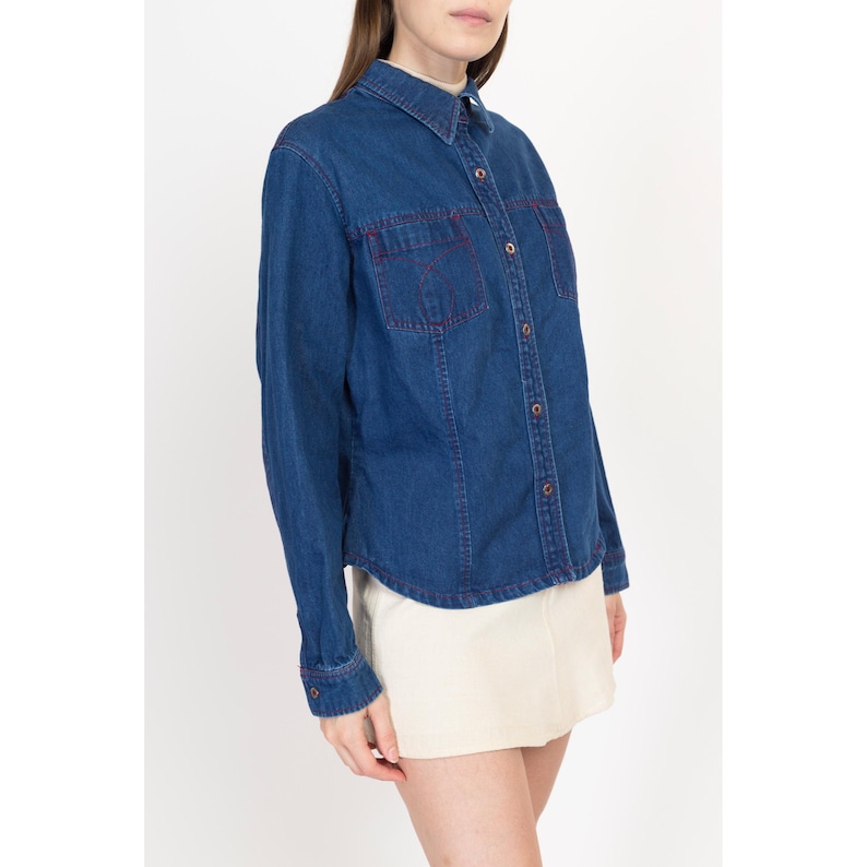 May include: A blue denim shirt with long sleeves and a button-up front. The shirt has red stitching around the pockets and collar.