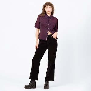 Puede incluir: Una mujer con una camisa morada oscura de manga corta y pantalones de pana negros. La camisa tiene cuello y un bolsillo. Los pantalones son de pierna ancha y la mujer lleva botas de plataforma.