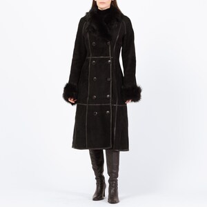 May include: A long, black double-breasted coat with a faux fur collar and cuffs. The coat features dark brown stitching and buttons. The model is wearing black boots. A classic winter coat.