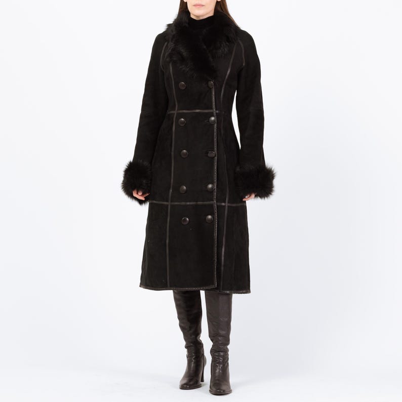 May include: A long, black double-breasted coat with a faux fur collar and cuffs. The coat features dark brown stitching and buttons. The model is wearing black boots. A classic winter coat.