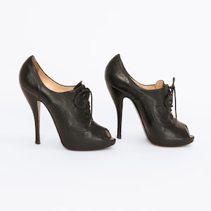 May include: A pair of black leather peep-toe high heels with lace-up detailing. The heels have a stiletto design with red soles. The shoes are set against a white background.