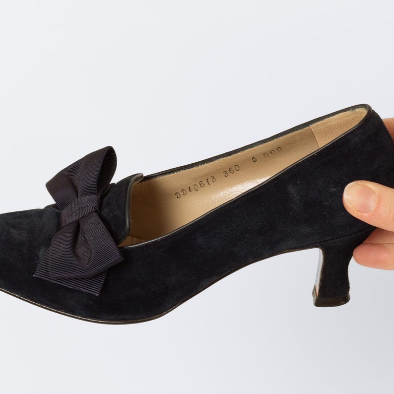 Puede incluir: Zapato negro de ante con tac&oacute;n bajo y detalle de lazo. El zapato tiene una puntera redondeada y un tac&oacute;n peque&ntilde;o. El lazo est&aacute; hecho de tela negra a juego. El zapato est&aacute; sostenido por una mano sobre un fondo blanco.
