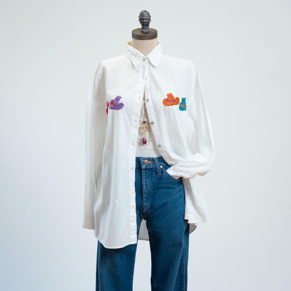 May include: A white long-sleeved button-down shirt featuring small, colorful embroidered shapes on the chest, worn over blue denim jeans. The shirt has a classic collar and a buttoned front placket, paired with a simple white undershirt.