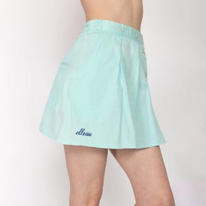 May include: A light blue skirt with a button-up front and a flared design. The skirt features the embroidered brand name "ellesse" in navy blue on the lower left side. The skirt is made of a lightweight fabric.