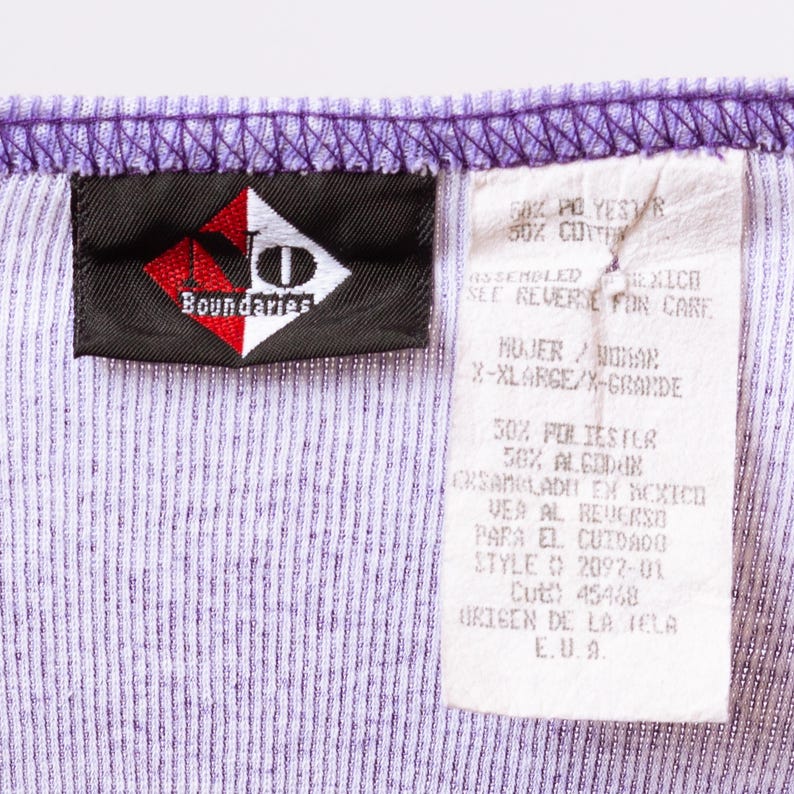 May include: A purple and white striped shirt tag with the brand name "Boundaries" and care instructions. The tag says "50% Polyester 50% Cotton" and "Assembled in Mexico, See Reverse for Care". It also says "Mujer/Woman, X-Large/X-Grande" and "50% Poli&eacute;ster 50% Algod&oacute;n, Ensamblado en M&eacute;xico, Vea al Reverso para el cuidado".