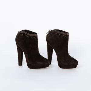 May include: A pair of dark brown suede ankle boots with high heels. The boots have a platform sole and a stitched design along the top edge. A zipper is visible on the back of one boot.