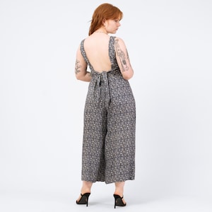 May include: A sleeveless jumpsuit with a low back and wide legs. The jumpsuit is navy blue with a small floral pattern in cream and beige. The back is tied with a bow. The model is wearing black high heels.
