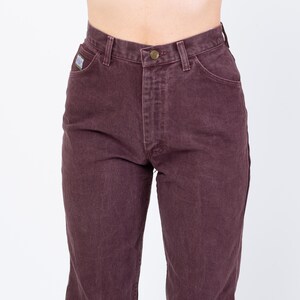 May include: Close-up of burgundy-colored denim jeans. The jeans have a button and zipper closure, classic five-pocket design, and a small logo patch on the front pocket. The jeans are a solid color and appear to be vintage.