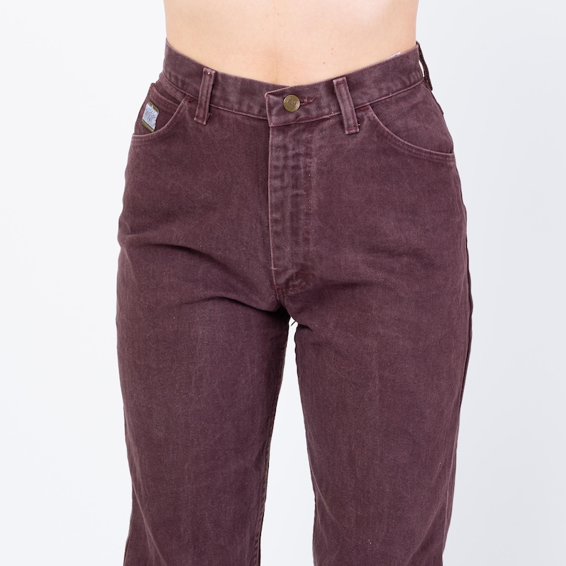 May include: Close-up of burgundy-colored denim jeans. The jeans have a button and zipper closure, classic five-pocket design, and a small logo patch on the front pocket. The jeans are a solid color and appear to be vintage.