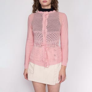 Puede incluir: Un cárdigan de punto rosa con un patrón de encaje y un lazo en la cintura.