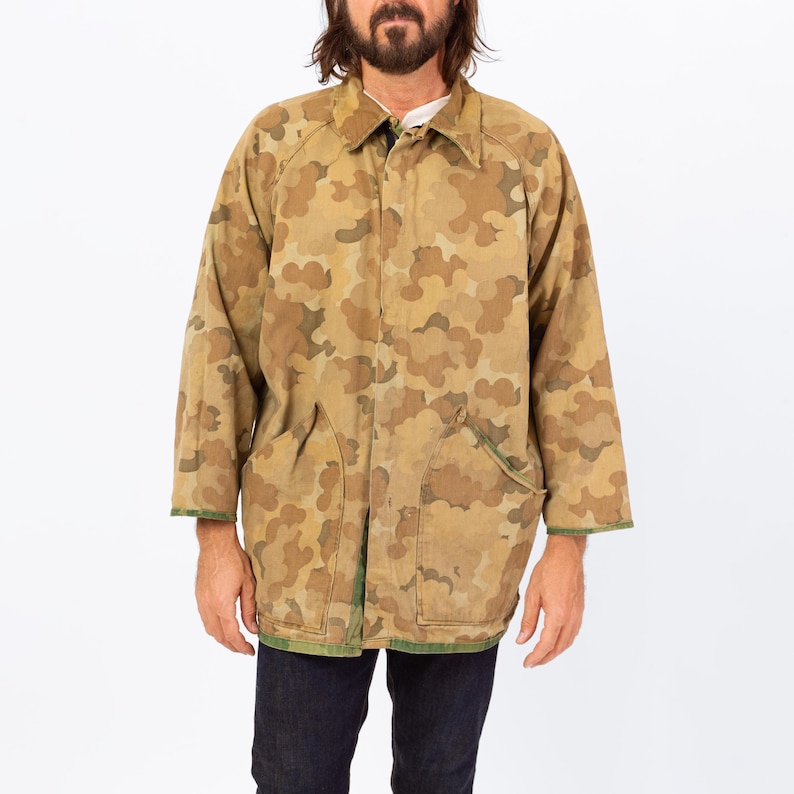 May include: A long, camouflage-patterned jacket in shades of brown and tan. The jacket has a collared neckline, two front pockets, and green trim along the cuffs and hem. The jacket is worn over a white shirt and dark blue jeans.