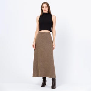 May include: A woman wearing a black sleeveless turtleneck top and a long, brown skirt. The skirt is a solid color and reaches the ankles. She is wearing black heeled boots. The outfit is simple and elegant.