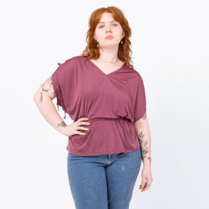 May include: A mauve-colored top with a V-neck and short, draped sleeves. The top has a cinched waist and is paired with blue jeans. The model is standing with one hand on their hip.