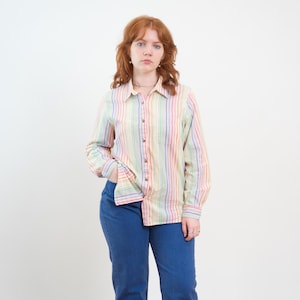 May include: A long-sleeved button-down shirt featuring a vertical striped pattern in pastel shades of yellow, pink, blue, and green. The shirt has a classic collar, a front button placket, and a single chest pocket, paired with solid blue denim jeans.