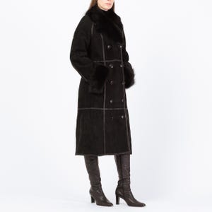 May include: A long, black double-breasted coat with a fur collar and cuffs. The coat features dark brown stitching and buttons. The person is wearing dark brown knee-high boots. The coat appears to be made of suede or a similar material.
