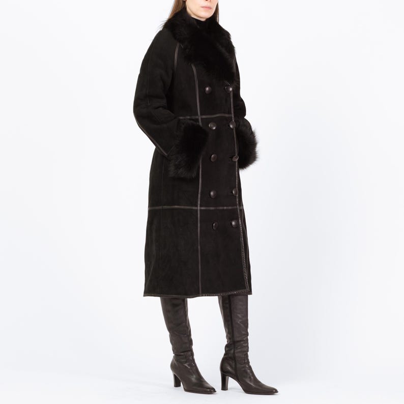 May include: A long, black double-breasted coat with a fur collar and cuffs. The coat features dark brown stitching and buttons. The person is wearing dark brown knee-high boots. The coat appears to be made of suede or a similar material.