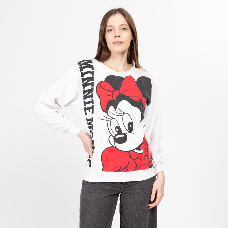 May include: White long-sleeve sweatshirt with a black and white image of Minnie Mouse wearing a red bow and red dress. The text "MINNIE MOUSE" is printed vertically on the left side of the image.