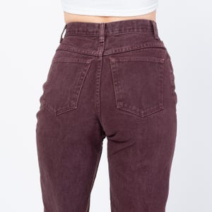 May include: Close-up of the back of a pair of high-waisted, burgundy-colored jeans. The jeans have two back pockets and a button and zipper closure. The fabric appears to be a sturdy denim material.