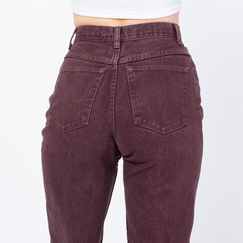 May include: Close-up of the back of a pair of high-waisted, burgundy-colored jeans. The jeans have two back pockets and a button and zipper closure. The fabric appears to be a sturdy denim material.