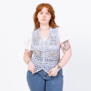 May include: A light blue, open-weave vest with floral embroidery. The vest has a button-up front and is worn over a white blouse with lace trim. The person is wearing blue jeans. The vest's design features a grid-like pattern.