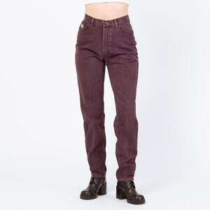 May include: A pair of high-waisted, plum-colored jeans. The jeans have a classic five-pocket design, a button and zipper closure, and a small logo patch on the back pocket. The model is wearing black heeled boots.