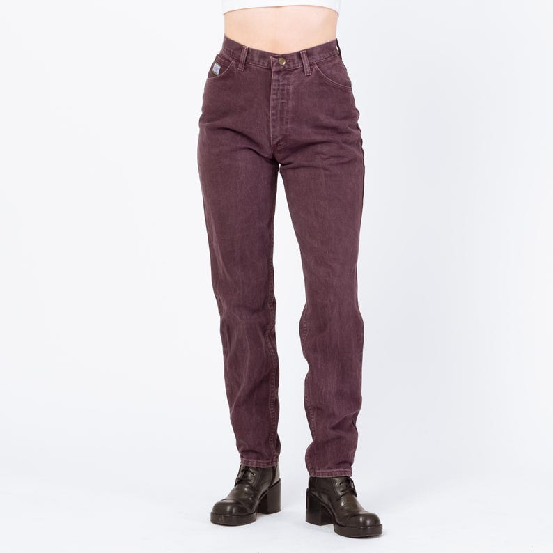 May include: A pair of high-waisted, plum-colored jeans. The jeans have a classic five-pocket design, a button and zipper closure, and a small logo patch on the back pocket. The model is wearing black heeled boots.