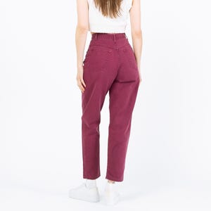 May include: A pair of high-waisted, burgundy-colored jeans. The jeans have a classic five-pocket design and a straight leg cut. The model is wearing a white top and white sneakers.