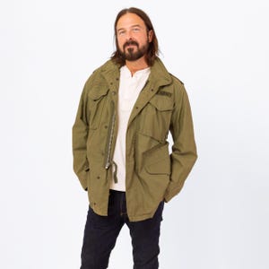 May include: Olive green military-style jacket with the word "ARMY" printed on the right chest. The jacket has multiple pockets, a zipper closure, and a drawstring at the waist. The person is wearing a white shirt and dark blue jeans.
